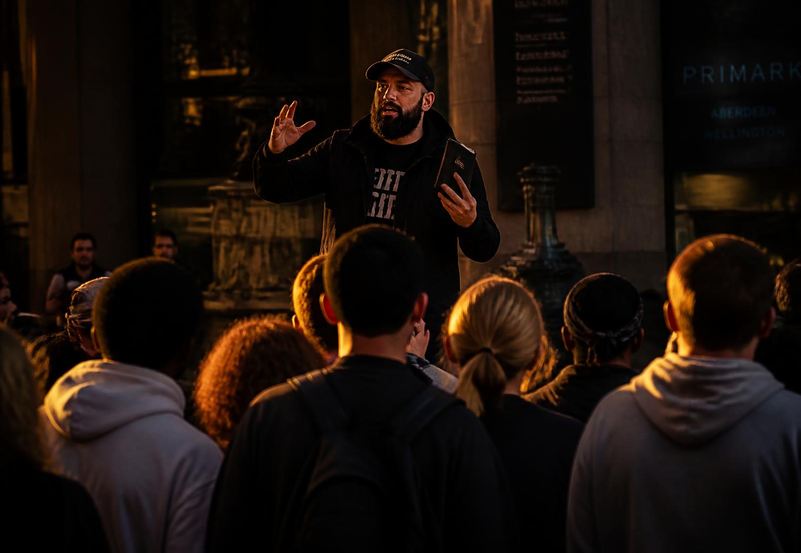 Hiubert Zamora preaching the gospel to a crowd gathered in a city plaza at dusk
